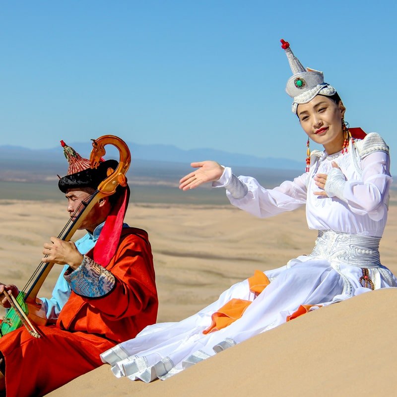 A scene in the Gobi desert featuring two individuals in traditional attire, one playing the Morin Khuur and the other a traditional dancer, sitting on the sand.