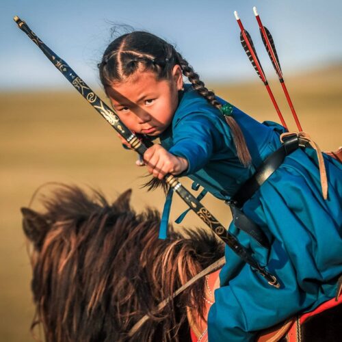 Young Mongolian girl loading an arrow on horseback.