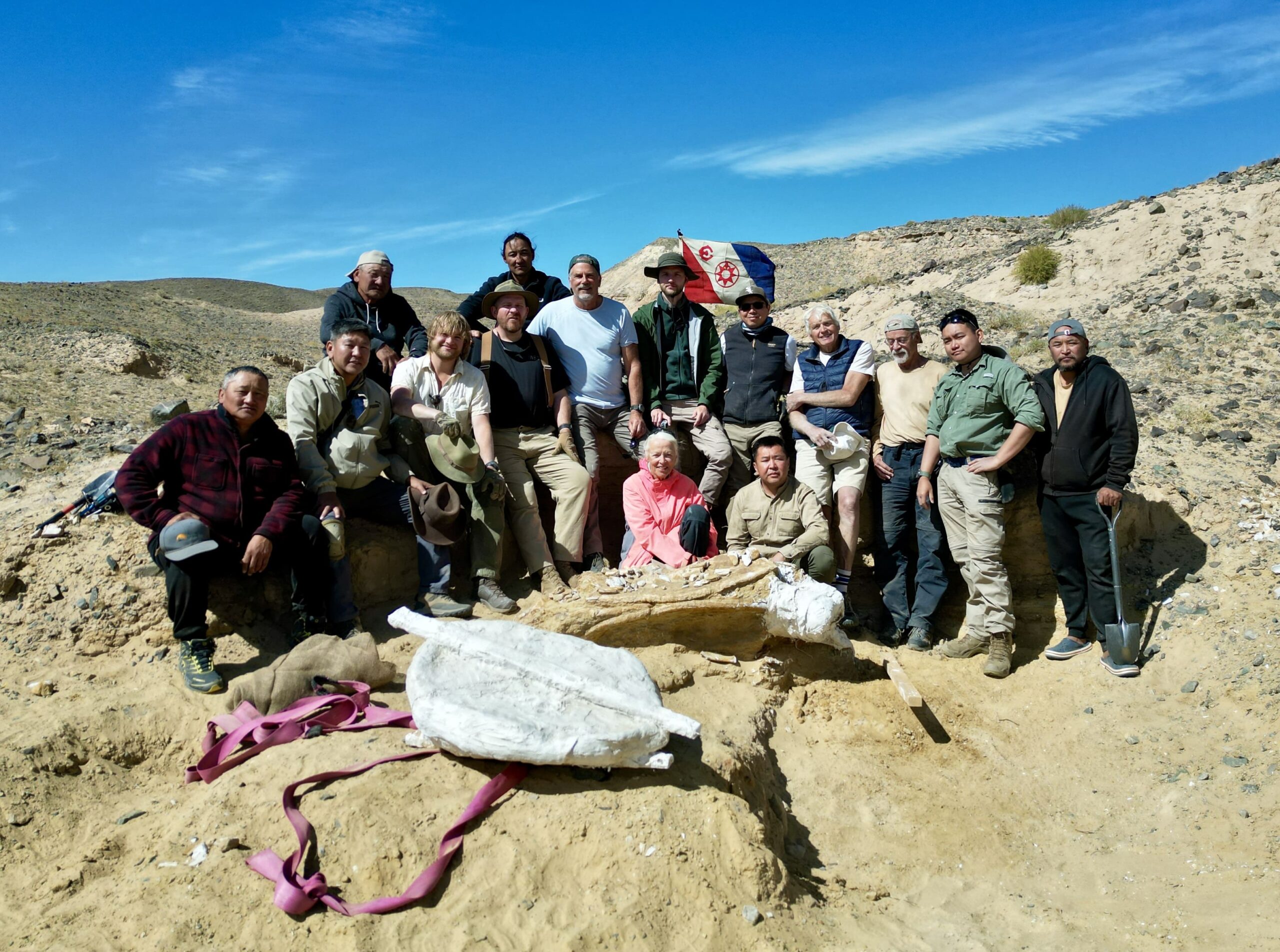 Group photo of the 2025 FLAG Expedition participants in the Gobi Desert, gathered beside a newly jacketed fossil with The Explorers Club flag displayed in the background.